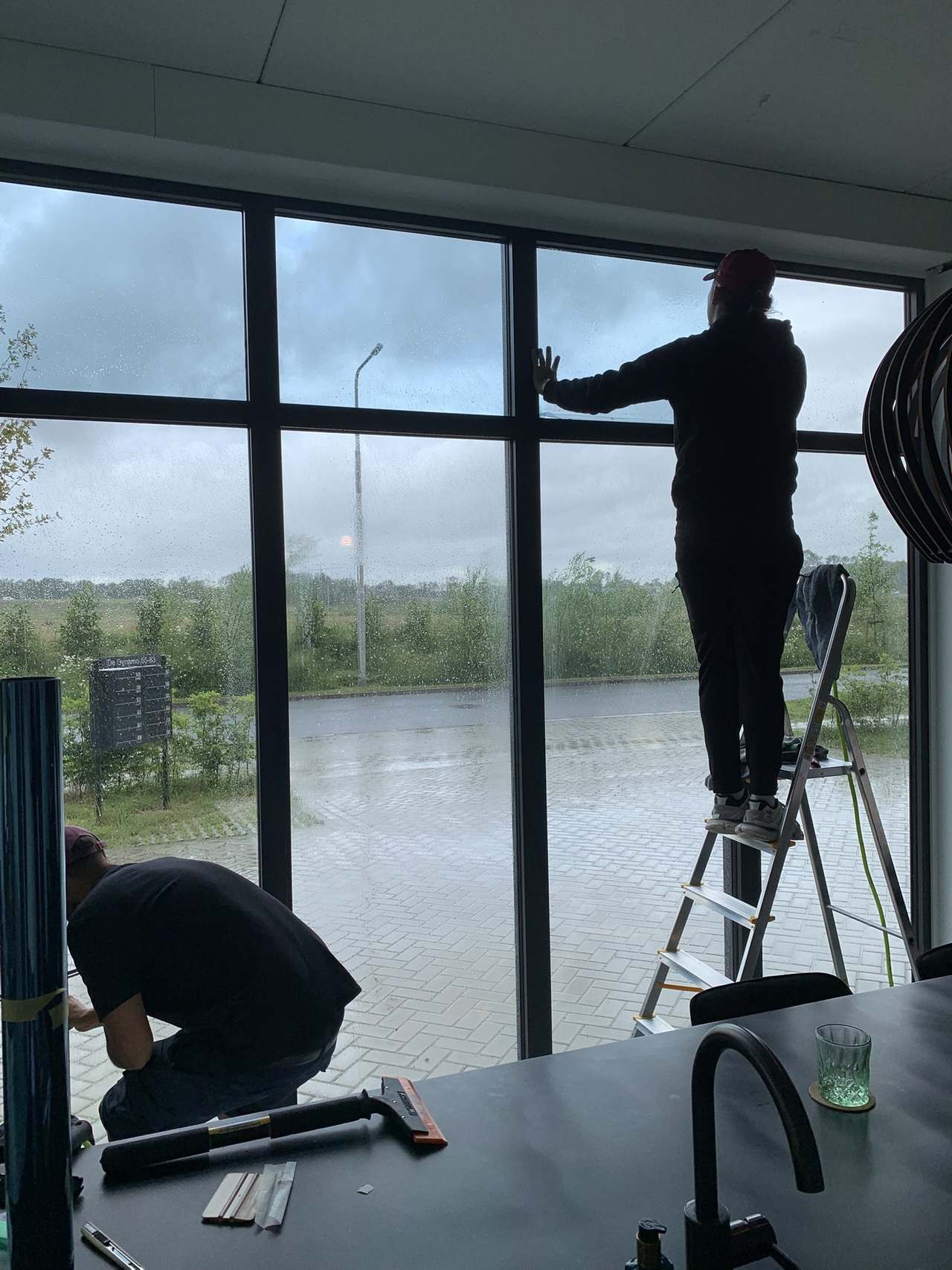 Twee monteurs brengen GlassShield raamfolie van binnenuit aan op een grote glazen pui, een van hen staat op een trapje